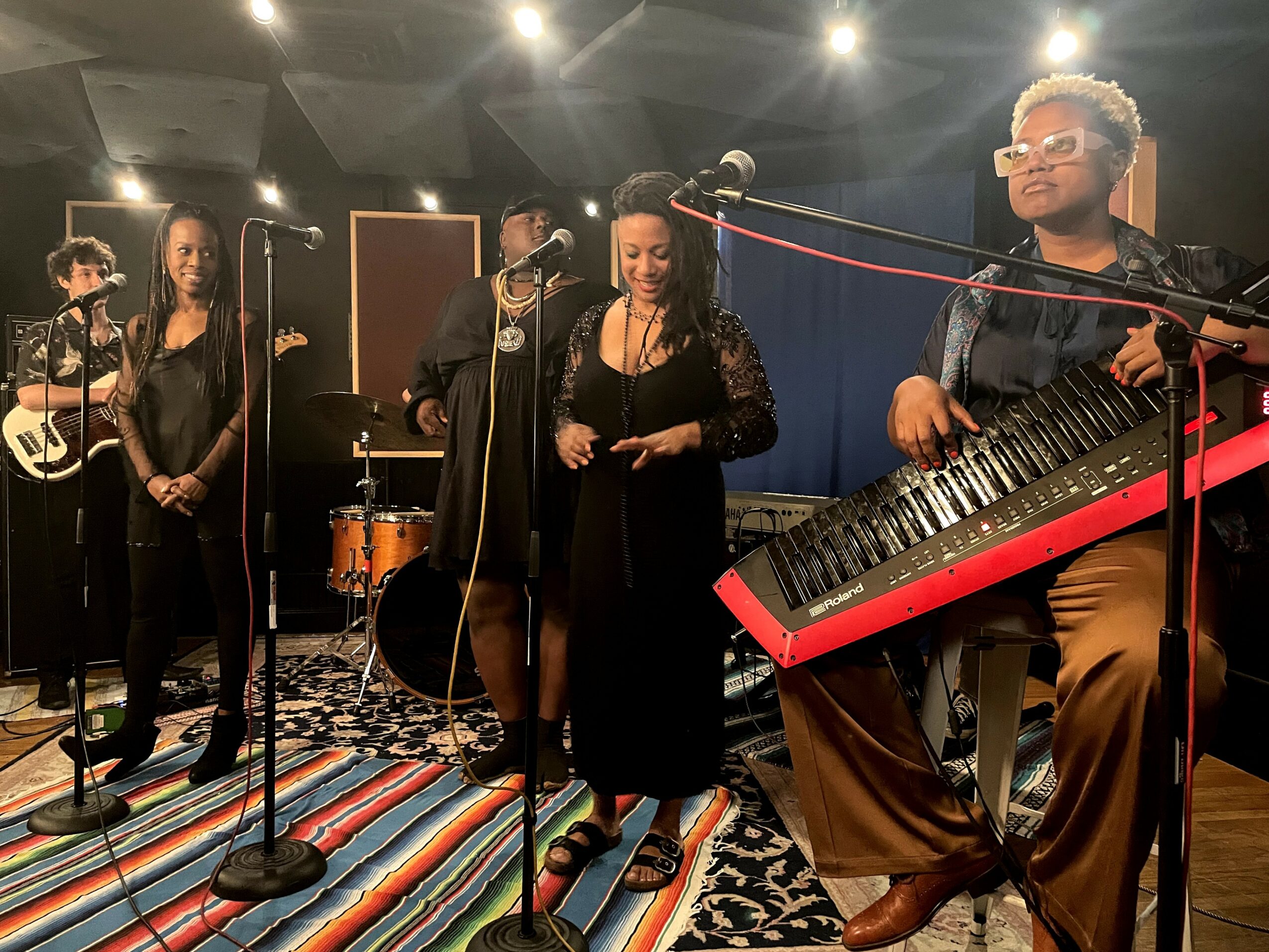 Group of five musicians performing on stage with instruments, colorful striped carpet, and dark background.