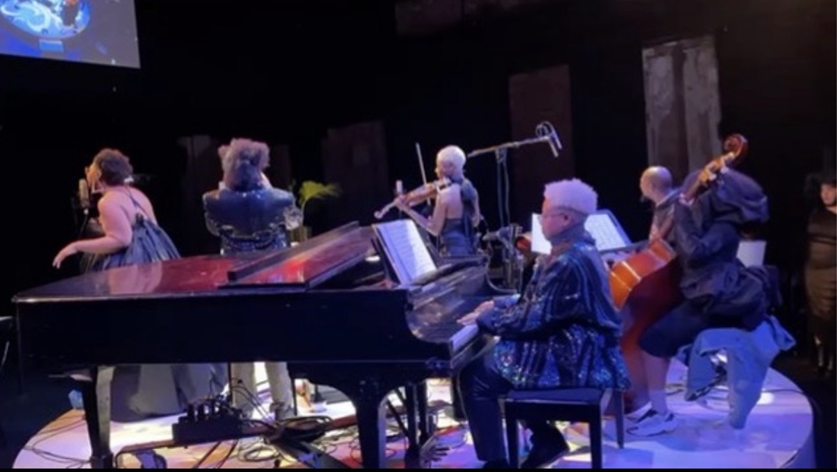 Group of musicians performing on stage with a grand piano and guitars, facing away from the camera.