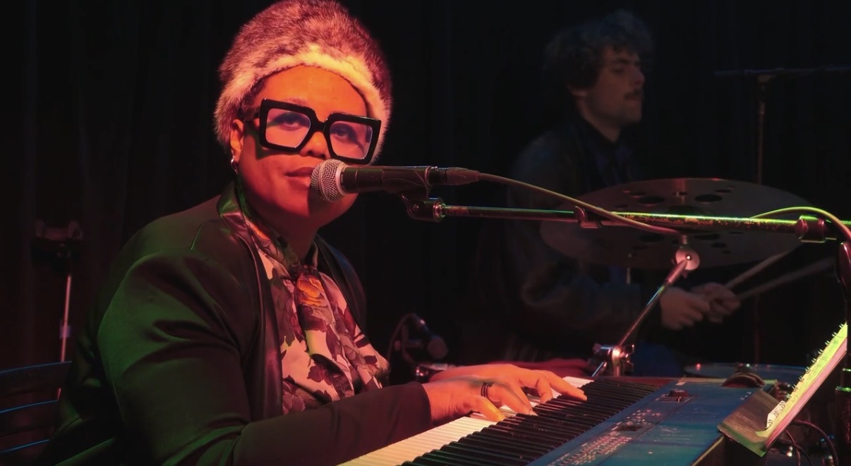 Person with glasses playing keyboard and singing into microphone on stage with a dark background.