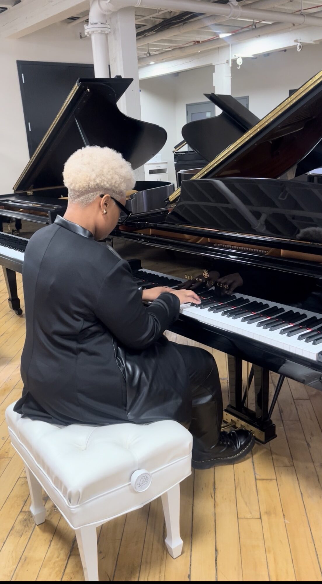 Person with curly hair playing a grand piano in a room with multiple pianos.