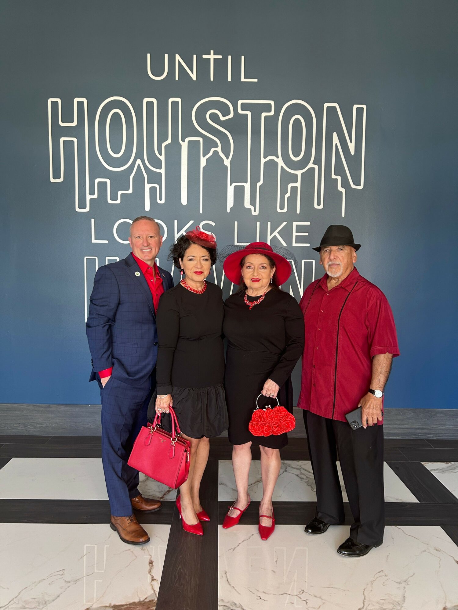 Four people standing in front of a wall with text, dressed in formal attire, holding handbags, smiling.