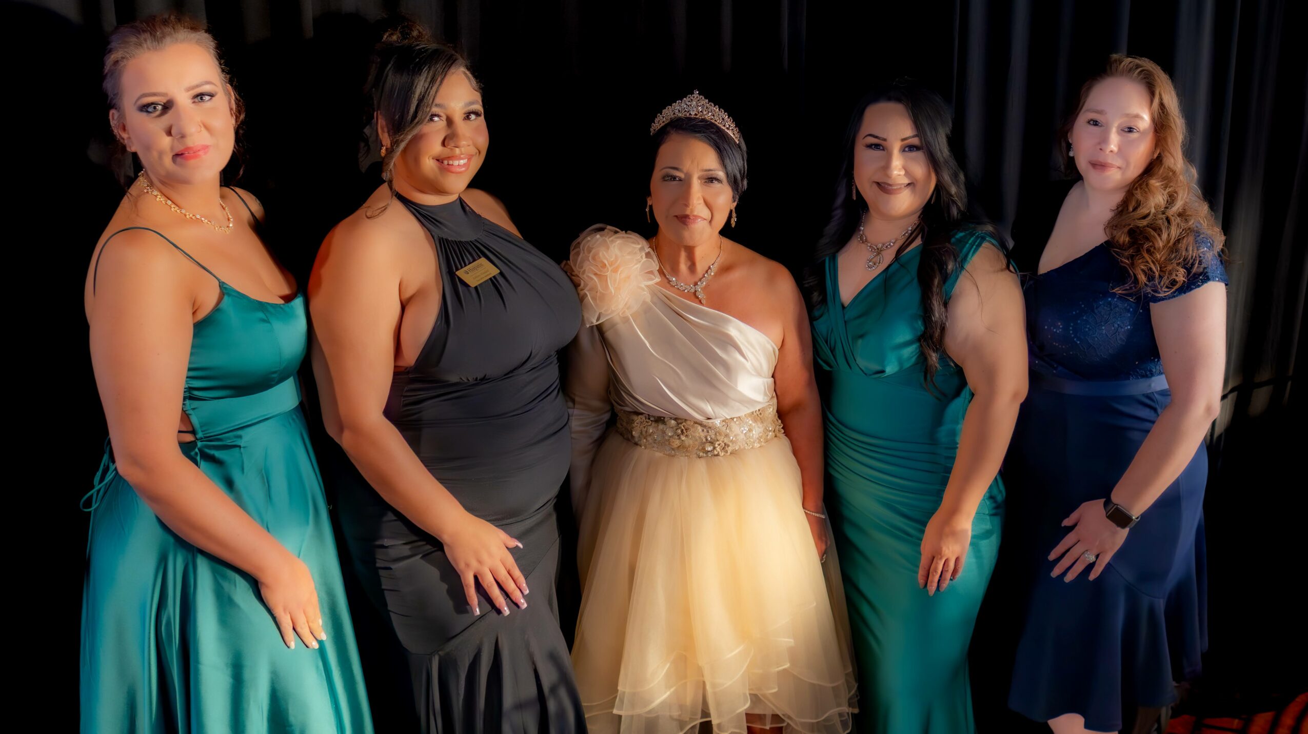 Five women in formal dresses standing together against a dark background.