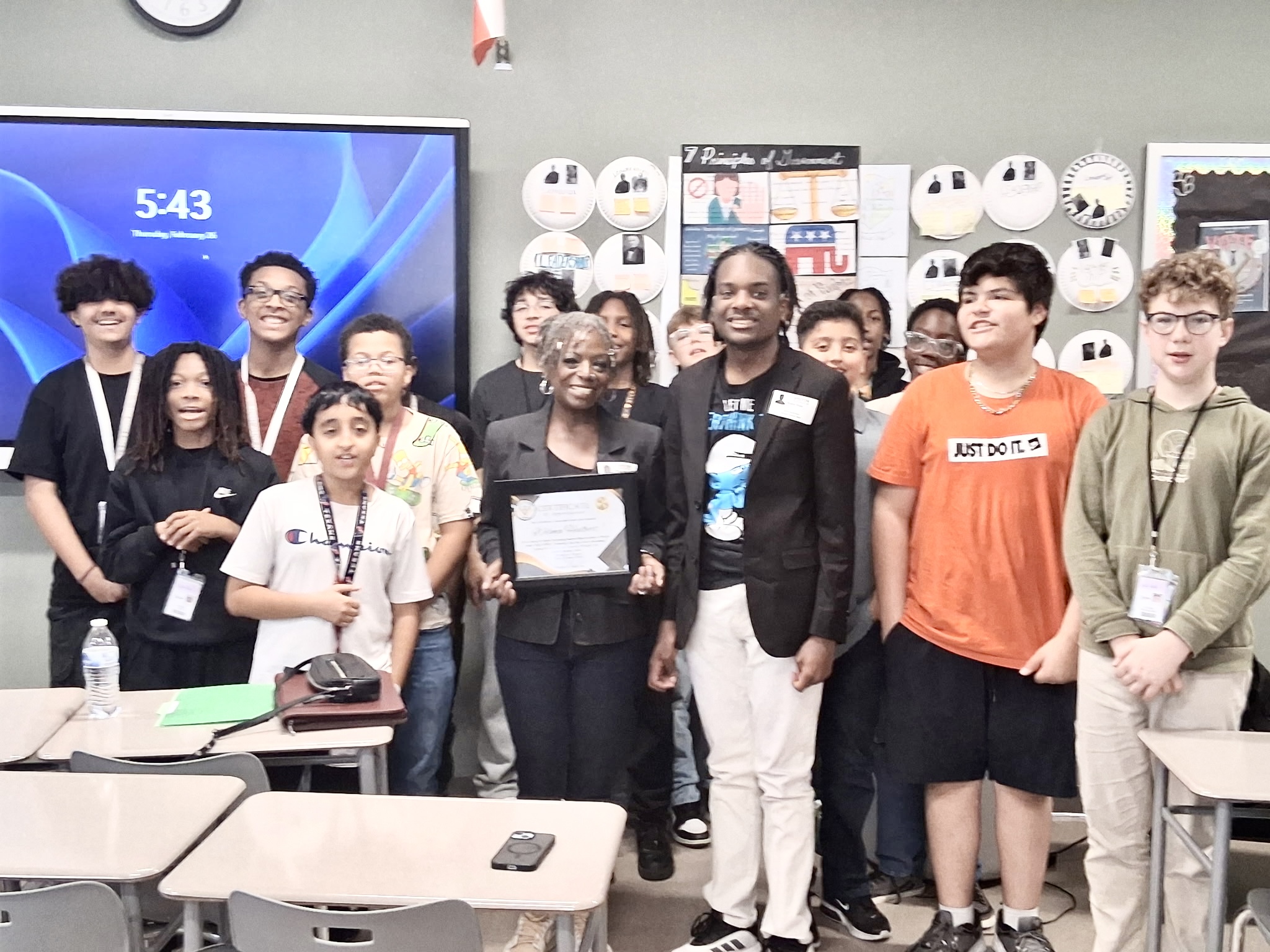 Group of diverse people standing in a classroom, some holding a framed certificate, with a screen displaying 5:43 in the background.