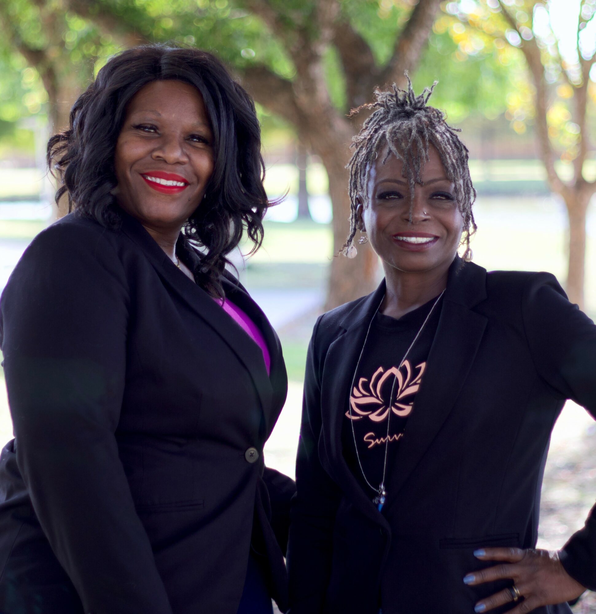 Two women smiling outdoors with trees in the background, wearing black blazers and casual tops.