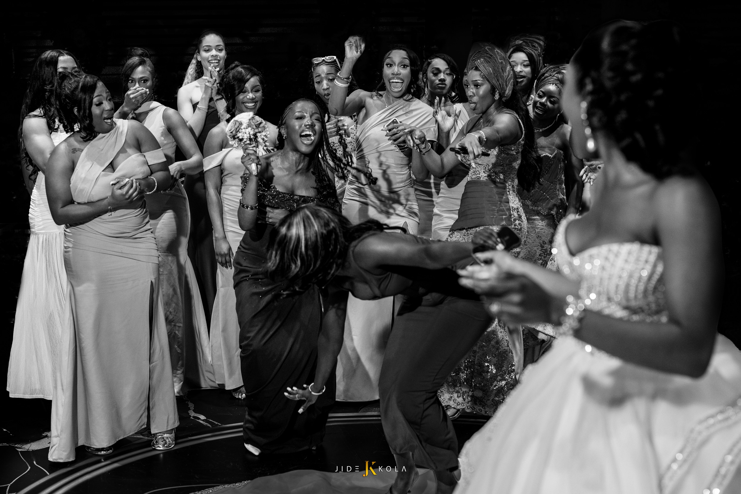 Group of women in formal dresses watching a woman perform a dance move, smiling and cheering, in a celebration setting.