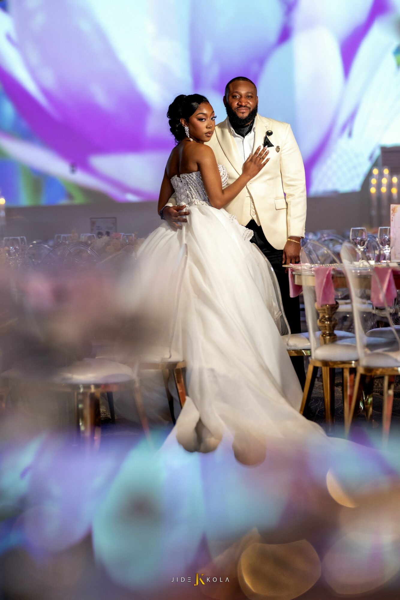 A bride and groom stand together at a wedding reception, with a large floral backdrop behind them. The bride wears a white gown, and the groom wears a white suit. They are holding a microphone and looking at each other. The reception area has decorated tables and chairs.