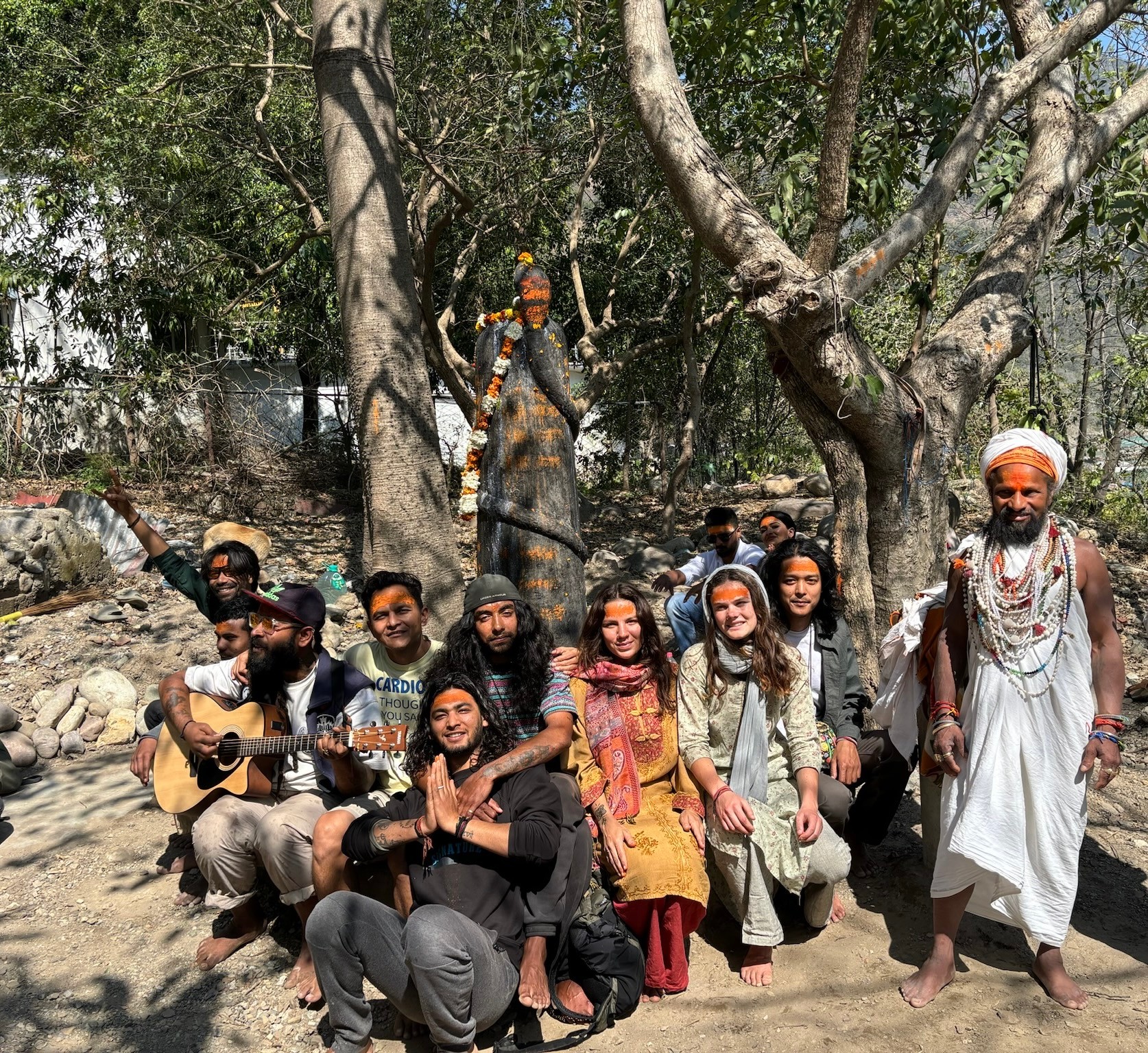 Group of people sitting and standing outdoors under trees, some with musical instruments, in a natural setting.
