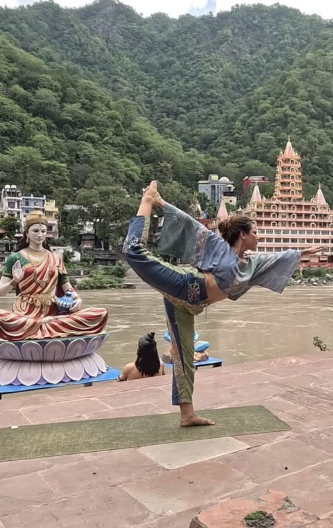 Person performing a yoga pose on a mat outdoors near water with a temple and green mountains in background.