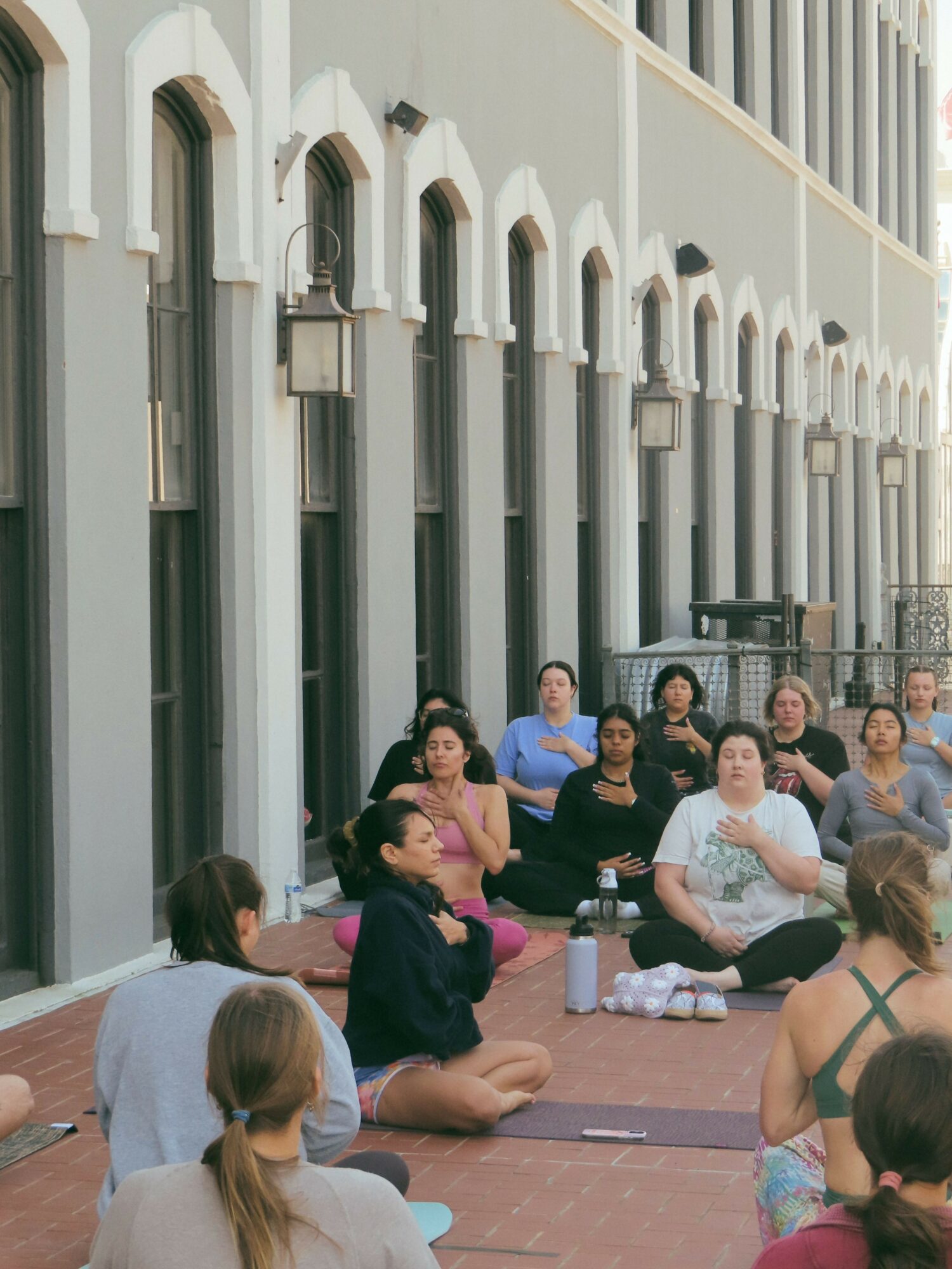 Group of people sitting on a brick floor outside a building with tall windows and decorative arches.
