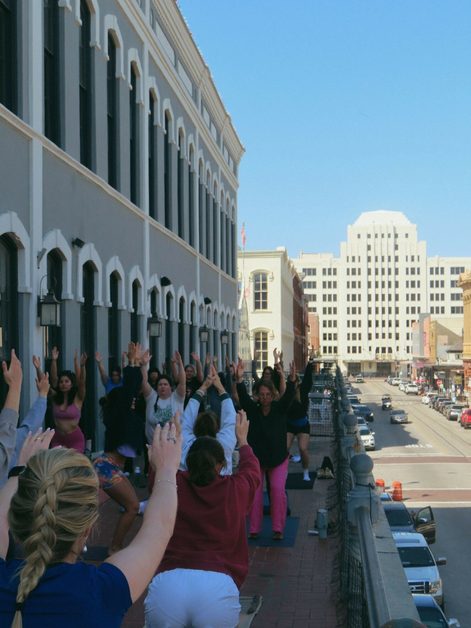 People walking on a city sidewalk with buildings in the background, some holding hands or talking, under a clear sky.