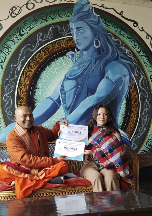 A man and woman sit in front of a large mural of a blue deity with multiple arms, holding certificates. The man wears orange robes, the woman a colorful shawl.