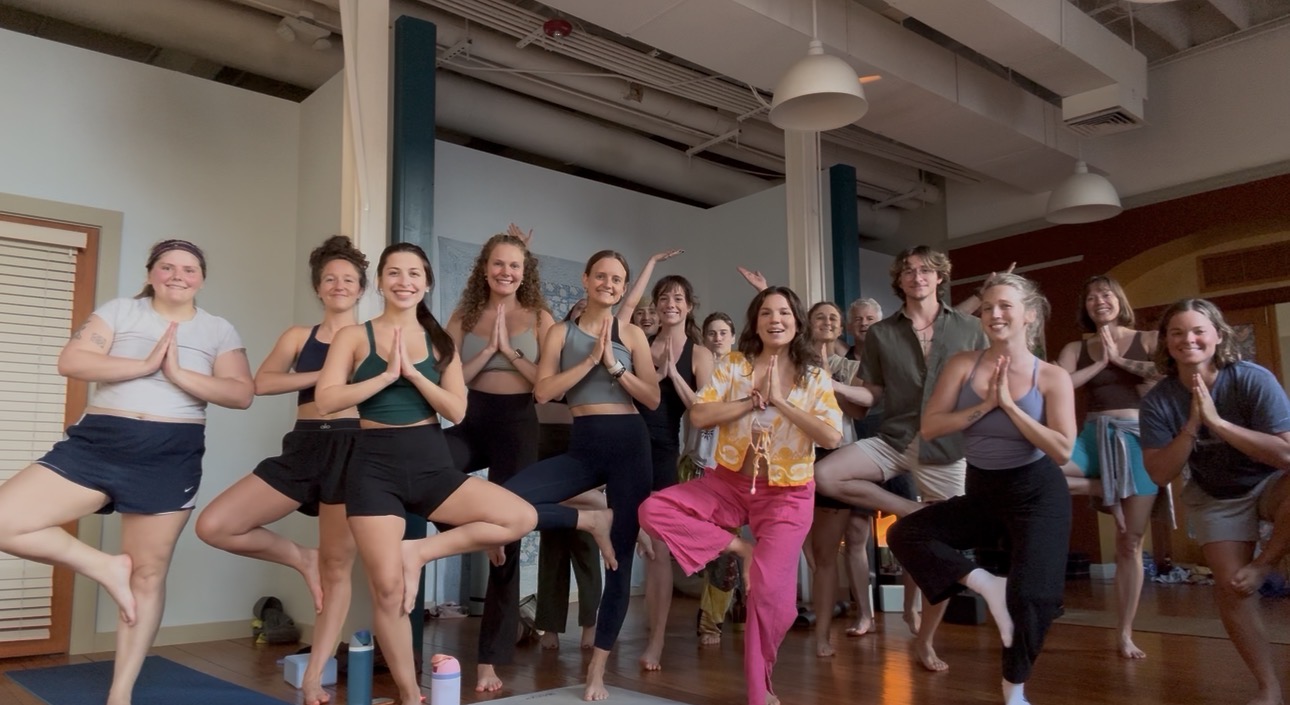 Group of people practicing yoga or meditation in a studio, some in tree pose, others sitting or standing, smiling.