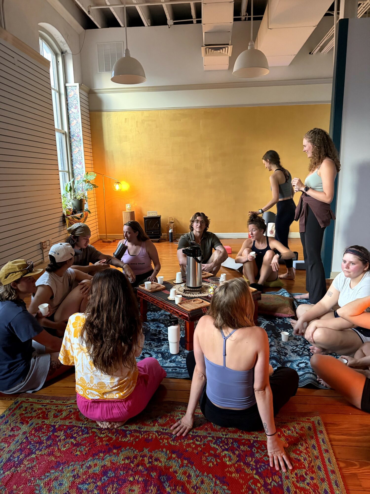 Group of people sitting on the floor and standing in a room with a yellow wall, large window, and wooden floor.