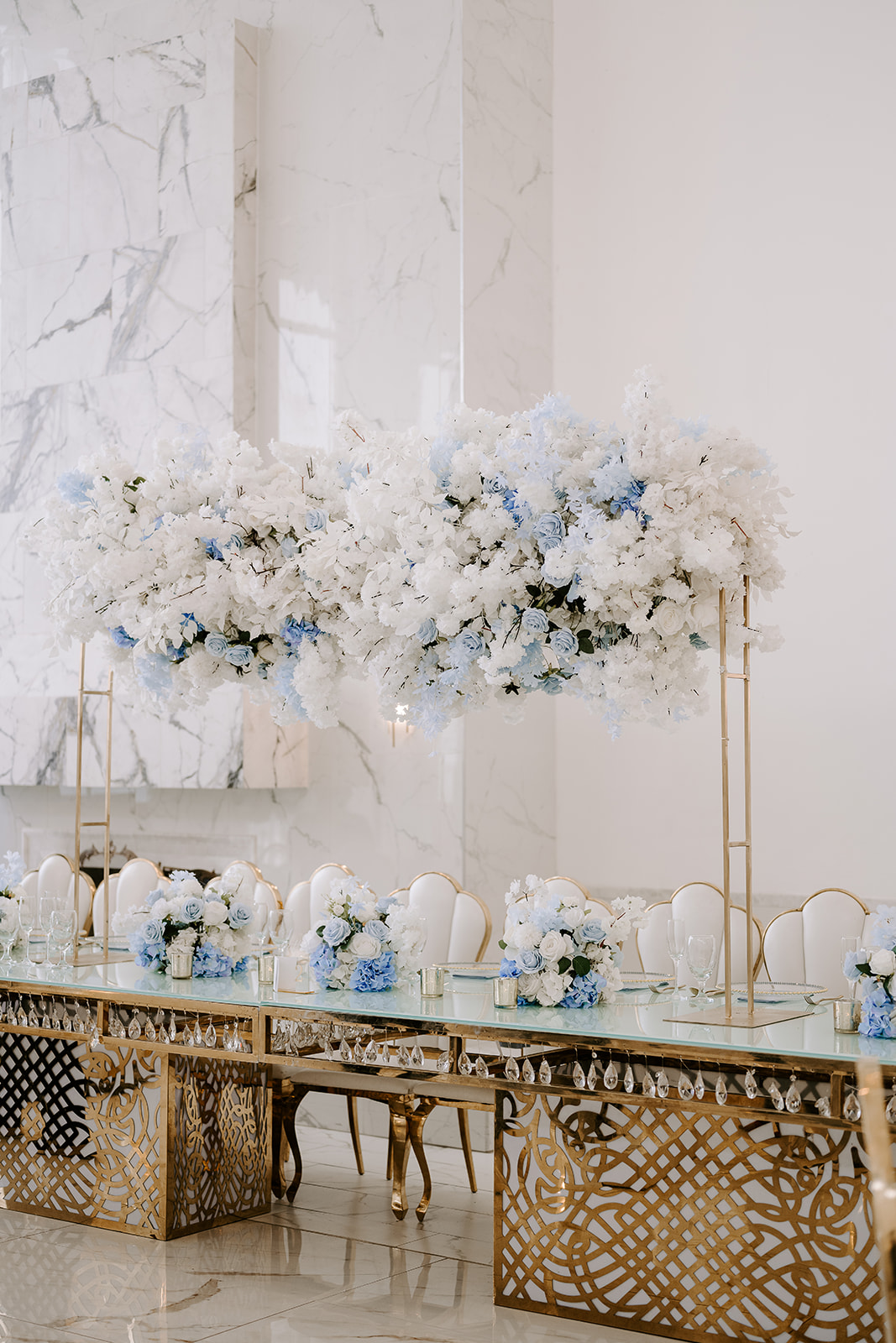 Elegant table with floral arrangements and chairs in a bright, white room, possibly for a wedding or event, with ornate gold accents.