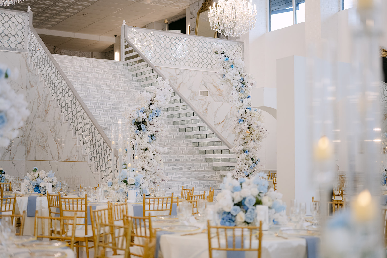 Elegant event space with white staircase decorated with flowers, gold chairs, and tall floral arrangements, bright natural light, and a chandelier.