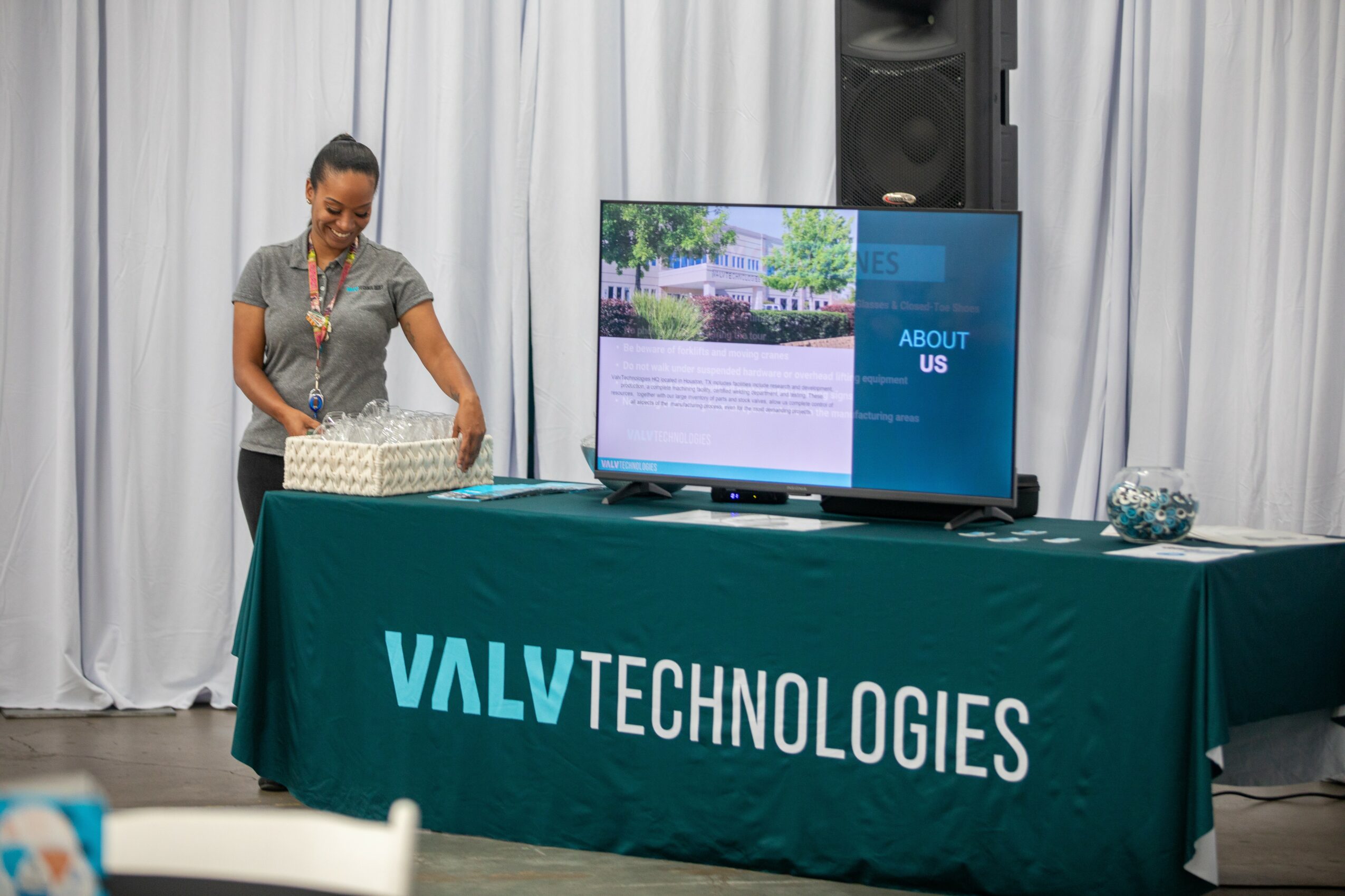 Woman standing behind a table with a VALV Technologies banner, a large screen displaying a presentation, and a white curtain backdrop.
