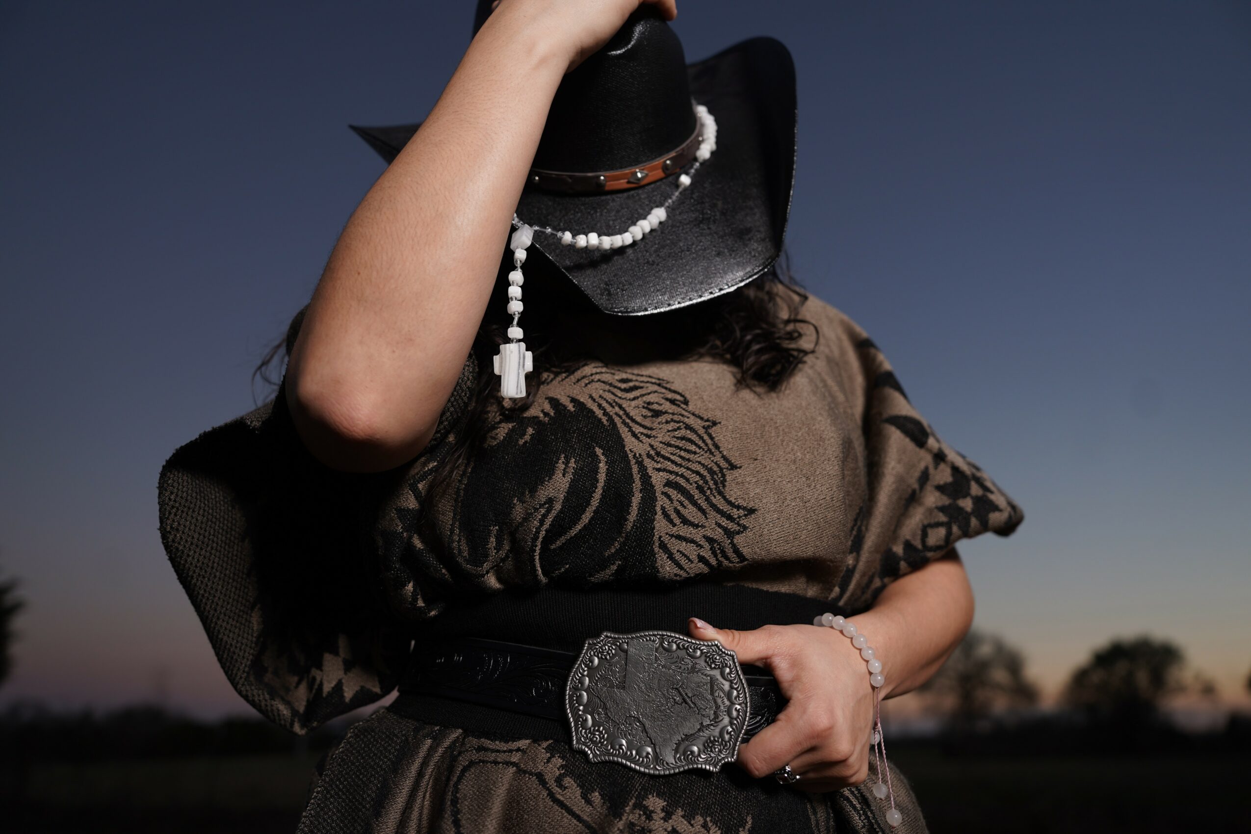 Person wearing a wide-brimmed hat, holding a large belt buckle, with a dark sky background.