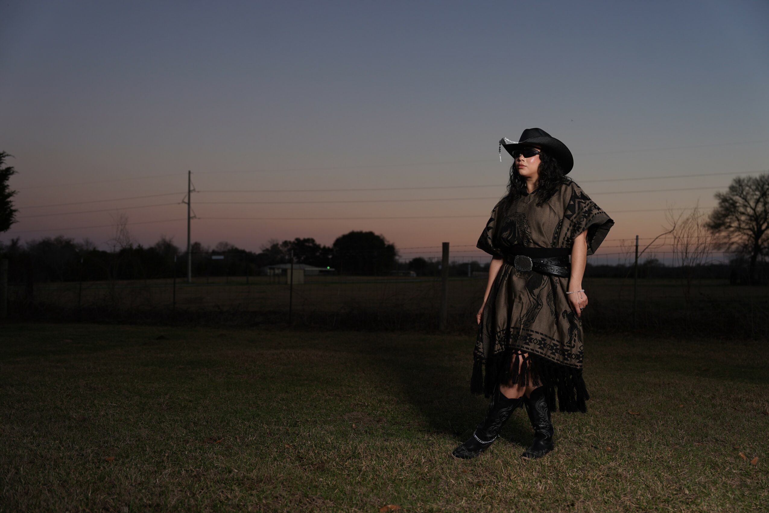 Person wearing a hat and dress standing outdoors during sunset, with trees and power lines in background.