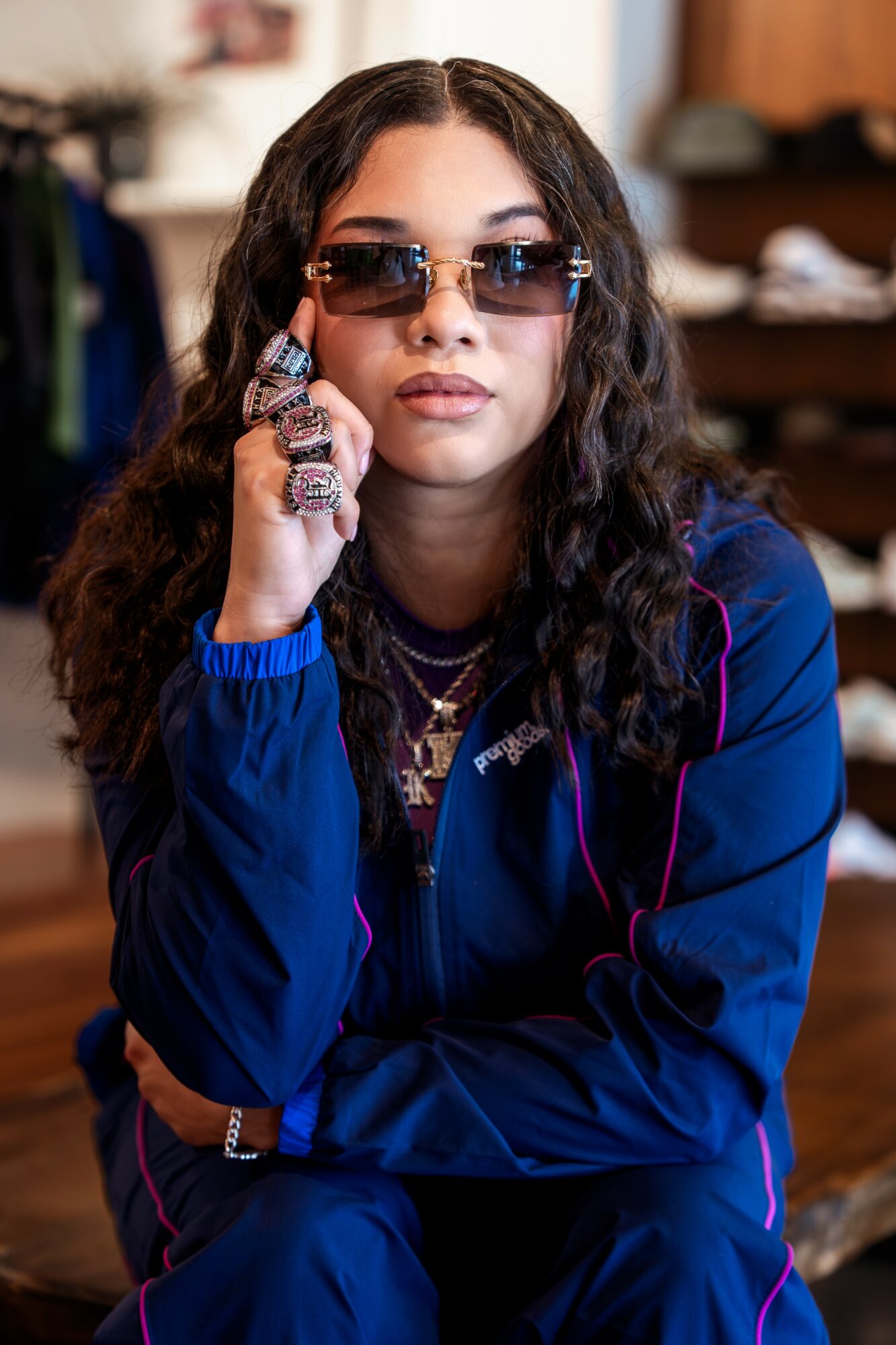 Young woman with curly hair wearing sunglasses and a blue jacket, sitting indoors with a serious expression.