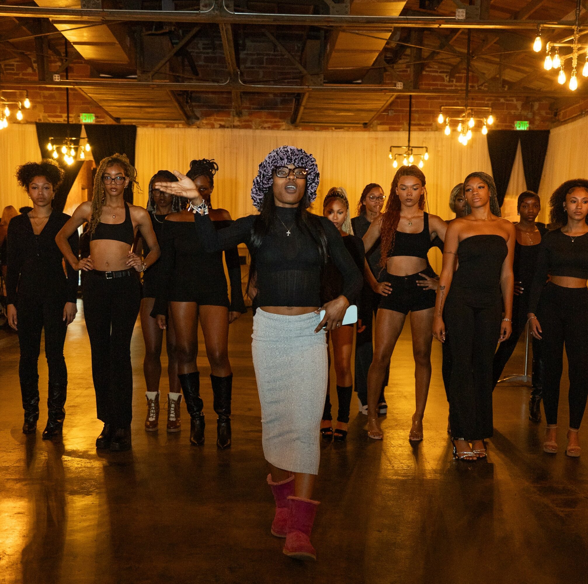 Group of women standing in a room with wooden floor and warm lighting, one woman in front wearing a hat and skirt.