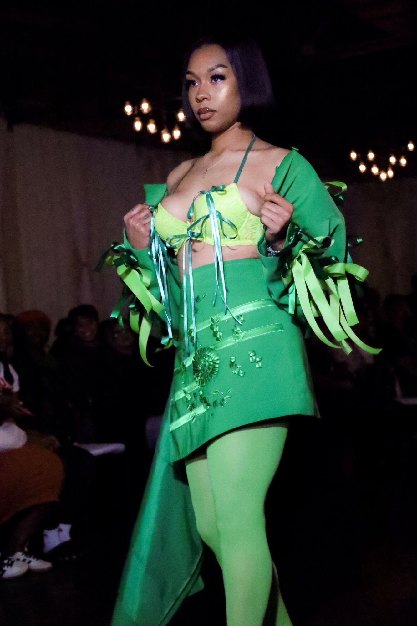 Model walking on runway in bright green outfit with ribbons and cutouts, dark background, audience visible, chandeliers overhead.