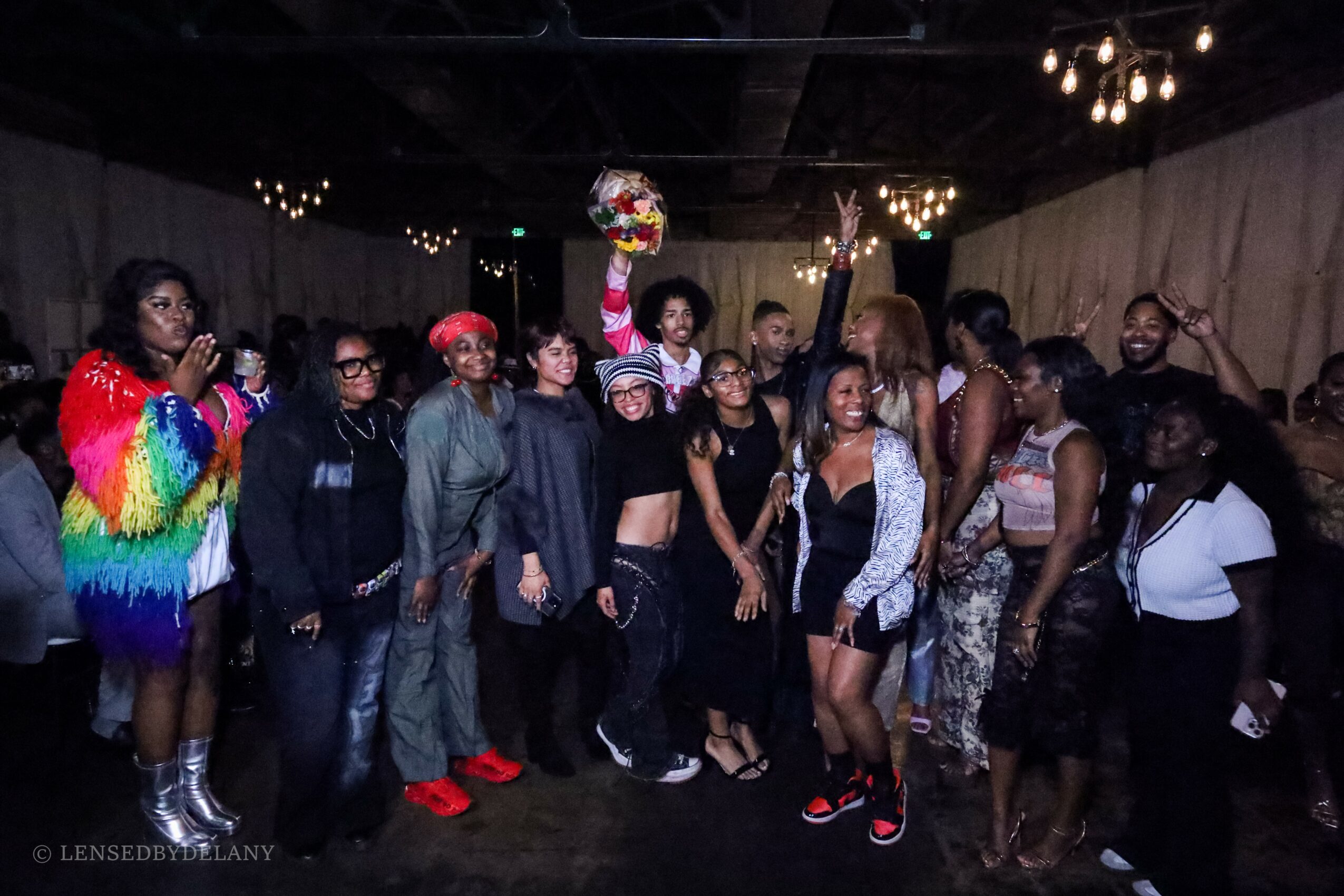 Group of people at a party, some dancing and others watching, with a person holding a bouquet in the air.