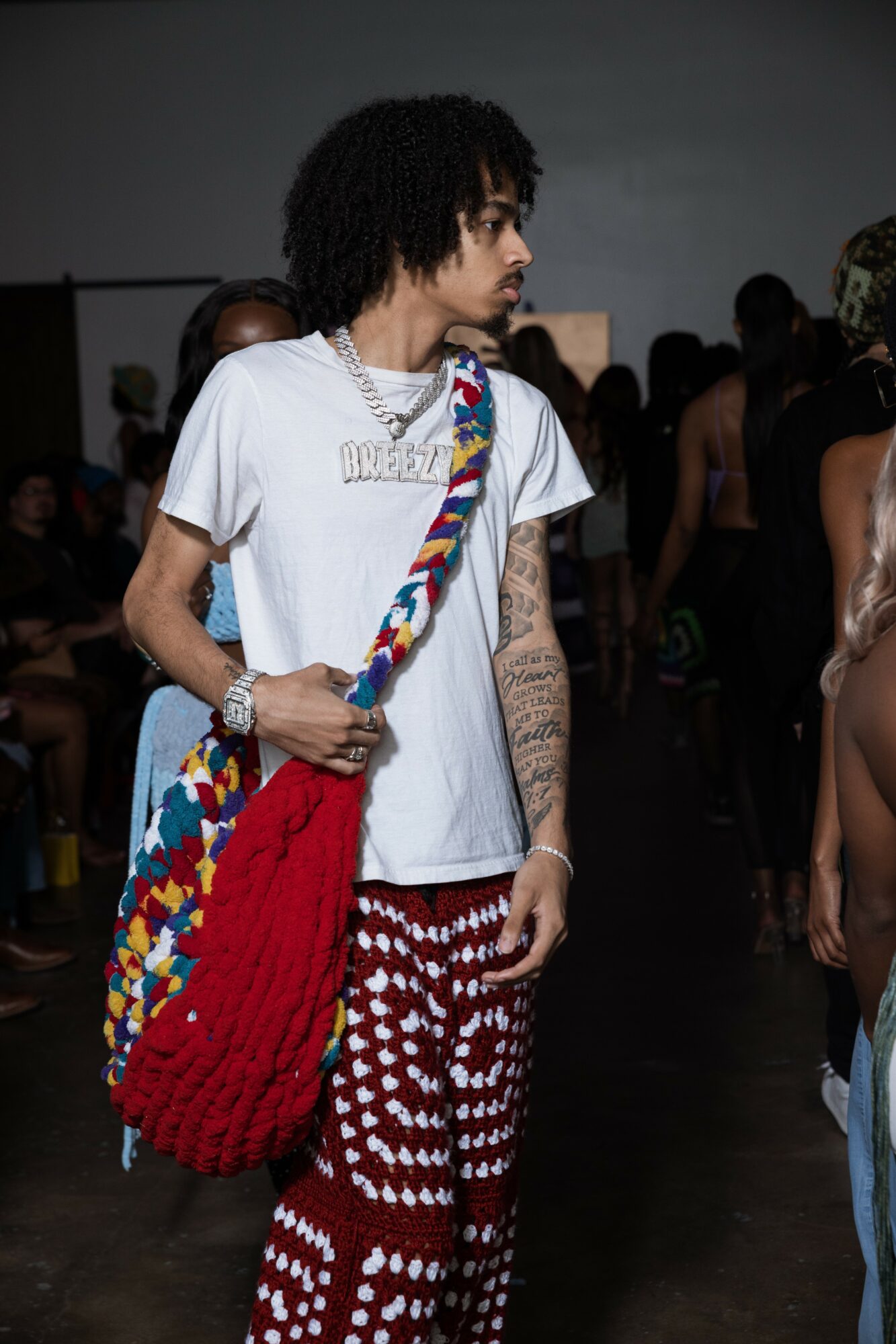 Person with curly hair wearing a white t-shirt, patterned pants, and carrying a colorful bag, in a crowded indoor space.