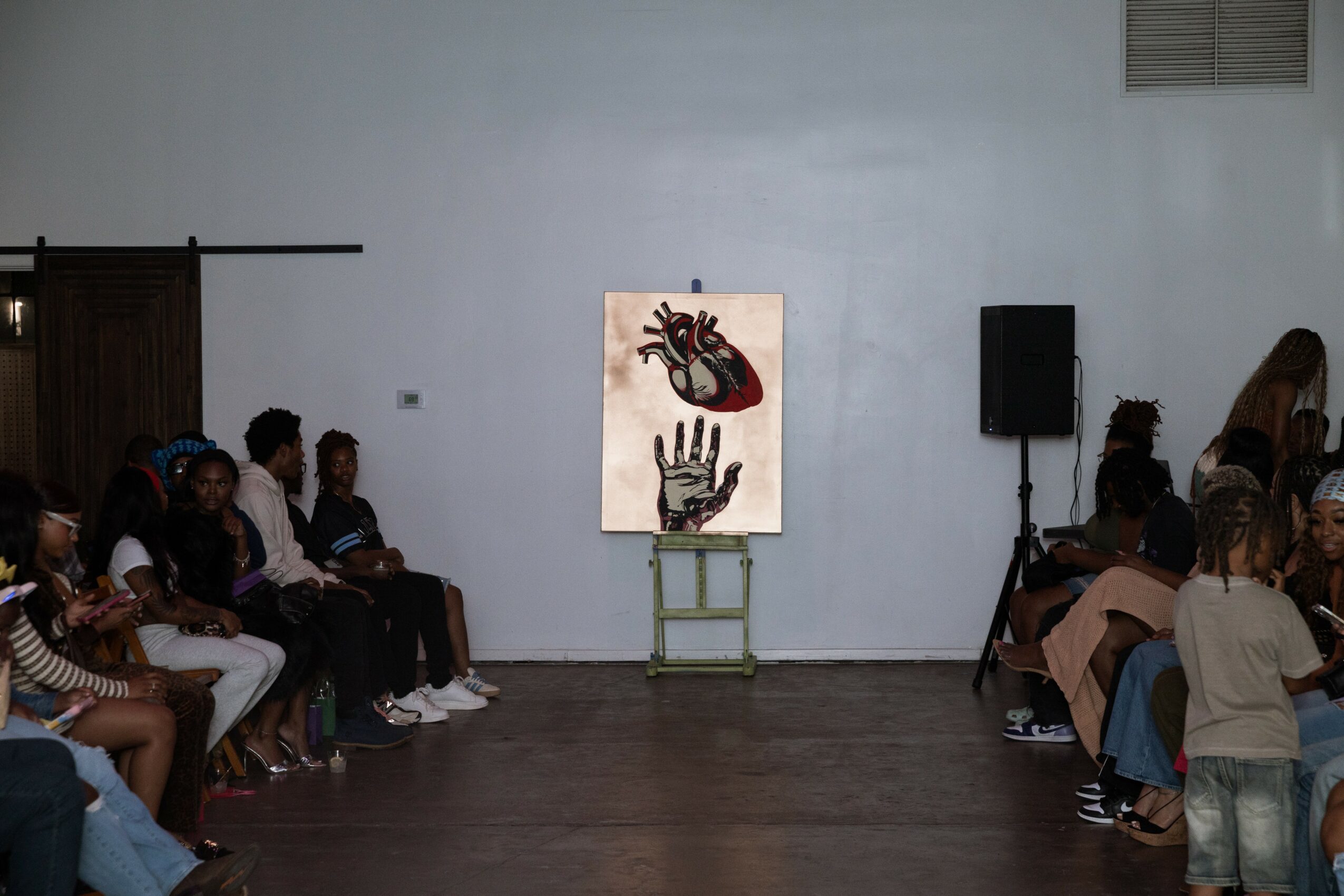 Audience seated on both sides facing a painting of a hand, a heart, and a face on a light blue wall.