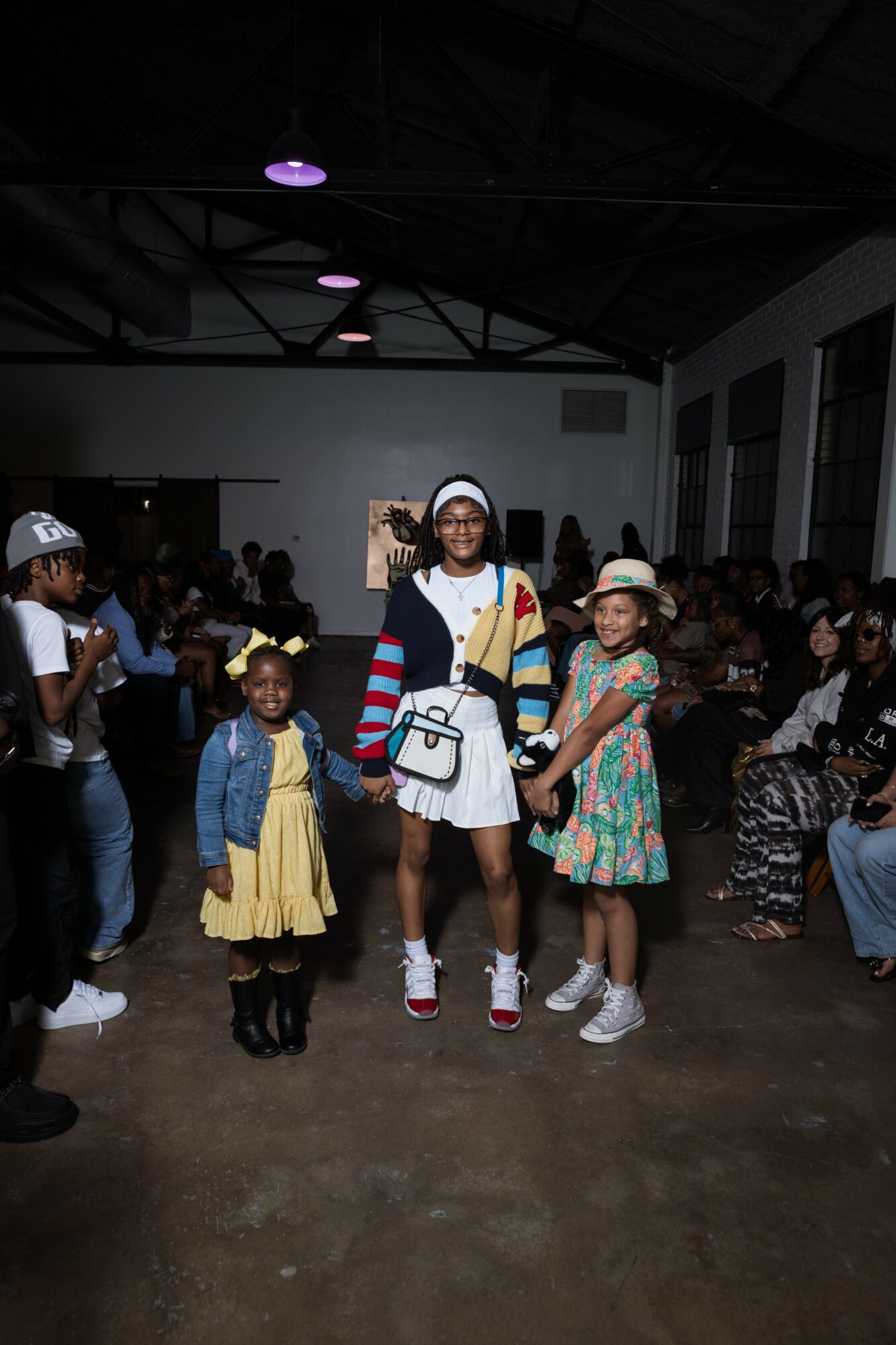 Three children and an adult standing together in a large indoor event space with seated audience in background.
