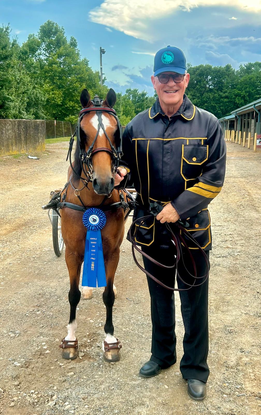 Person standing next to a horse with a blue ribbon, outdoors with trees and a building in the background.