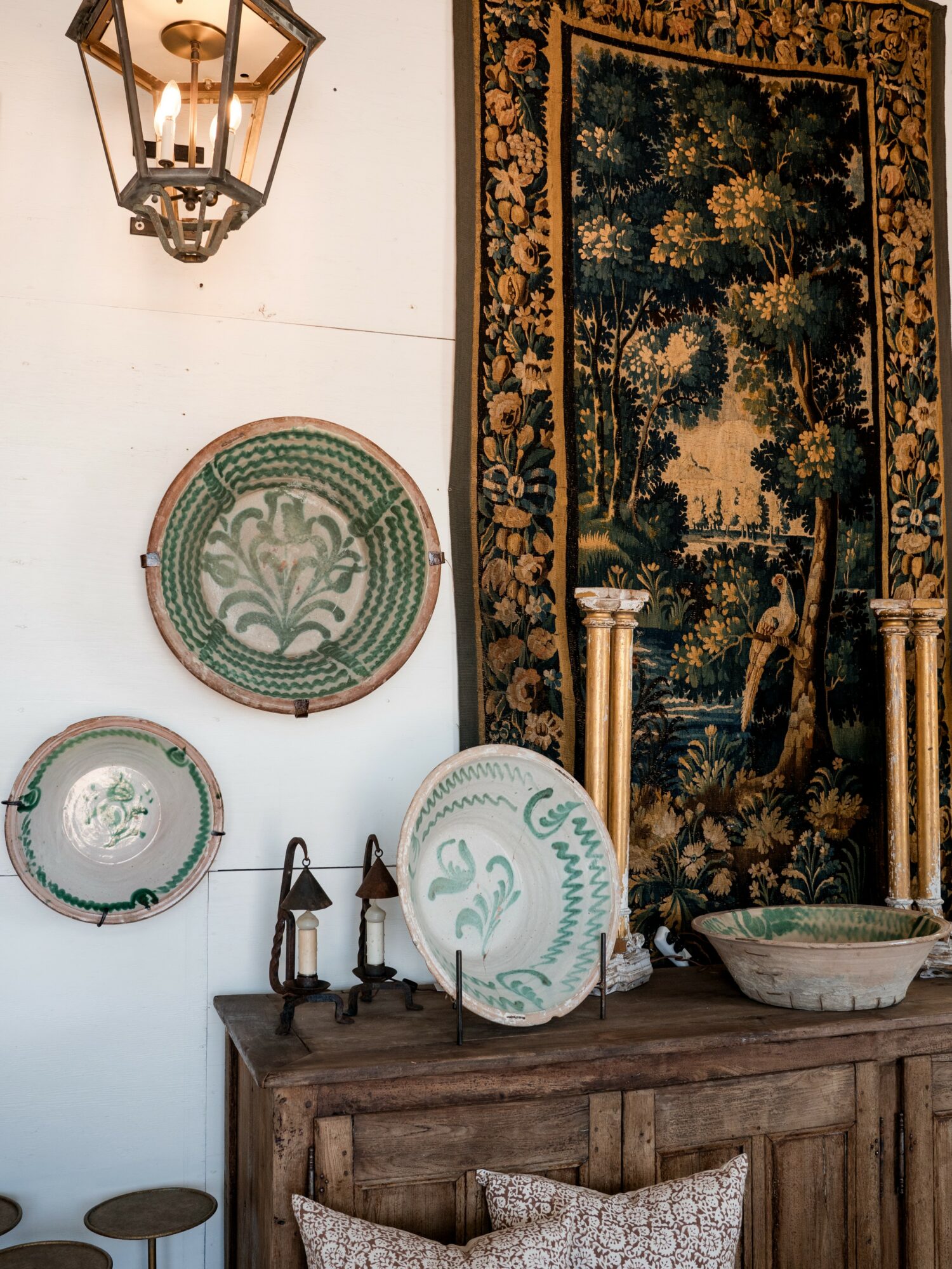 Decorative plates on a wall, a tapestry with trees, and a wooden cabinet with lamps and a bowl.