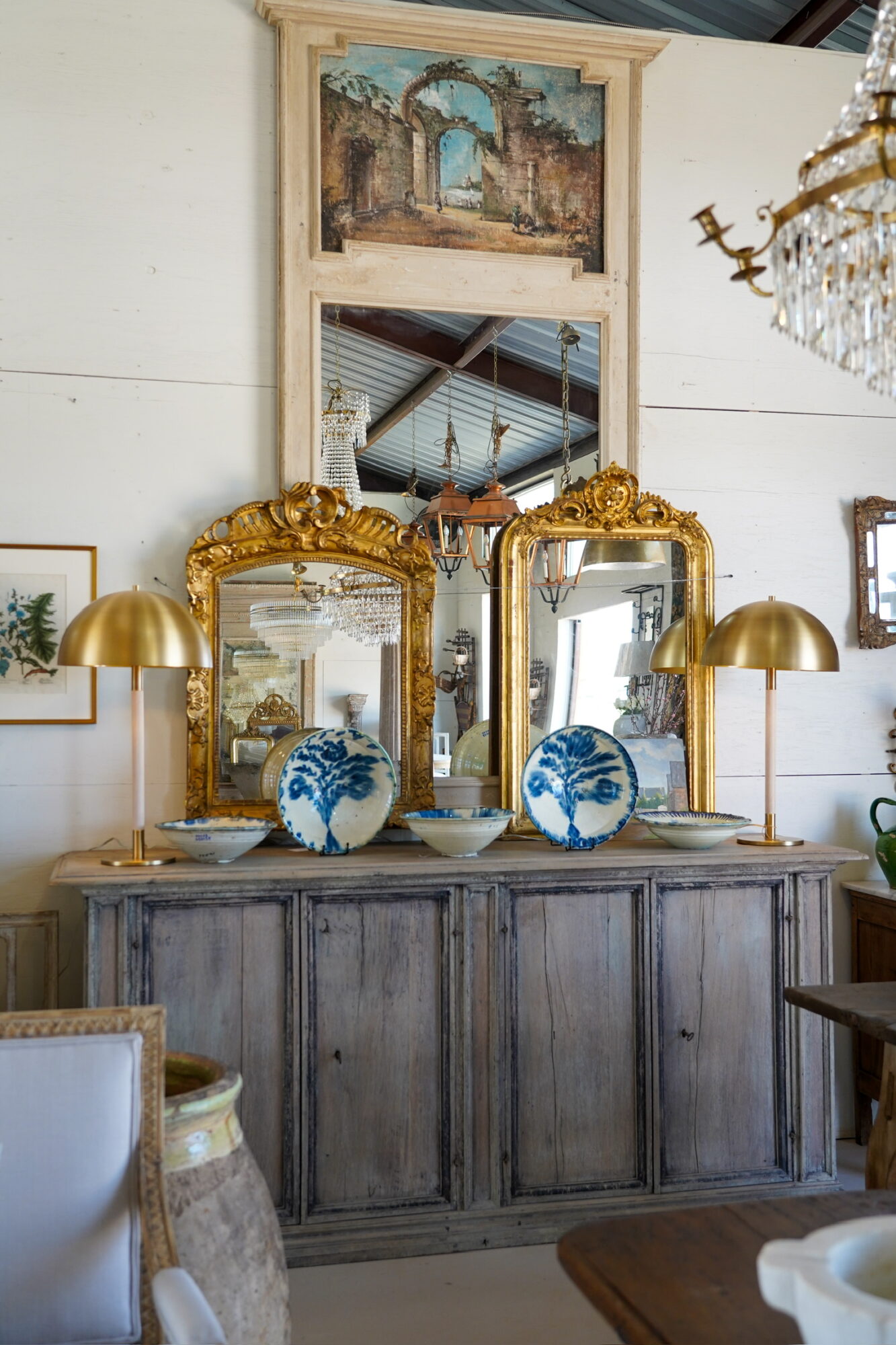 Decorative wooden sideboard with two ornate gold-framed mirrors, blue and white vases, and gold lamps, against a white wall with framed pictures and a chandelier.