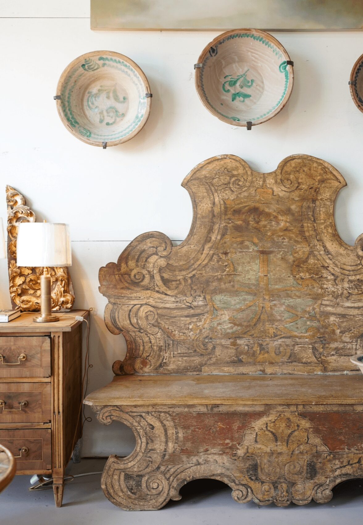 Decorative wooden headboard with intricate carvings, three painted ceramic plates on the wall, wooden nightstand with a lamp, and a gold-colored sculpture.