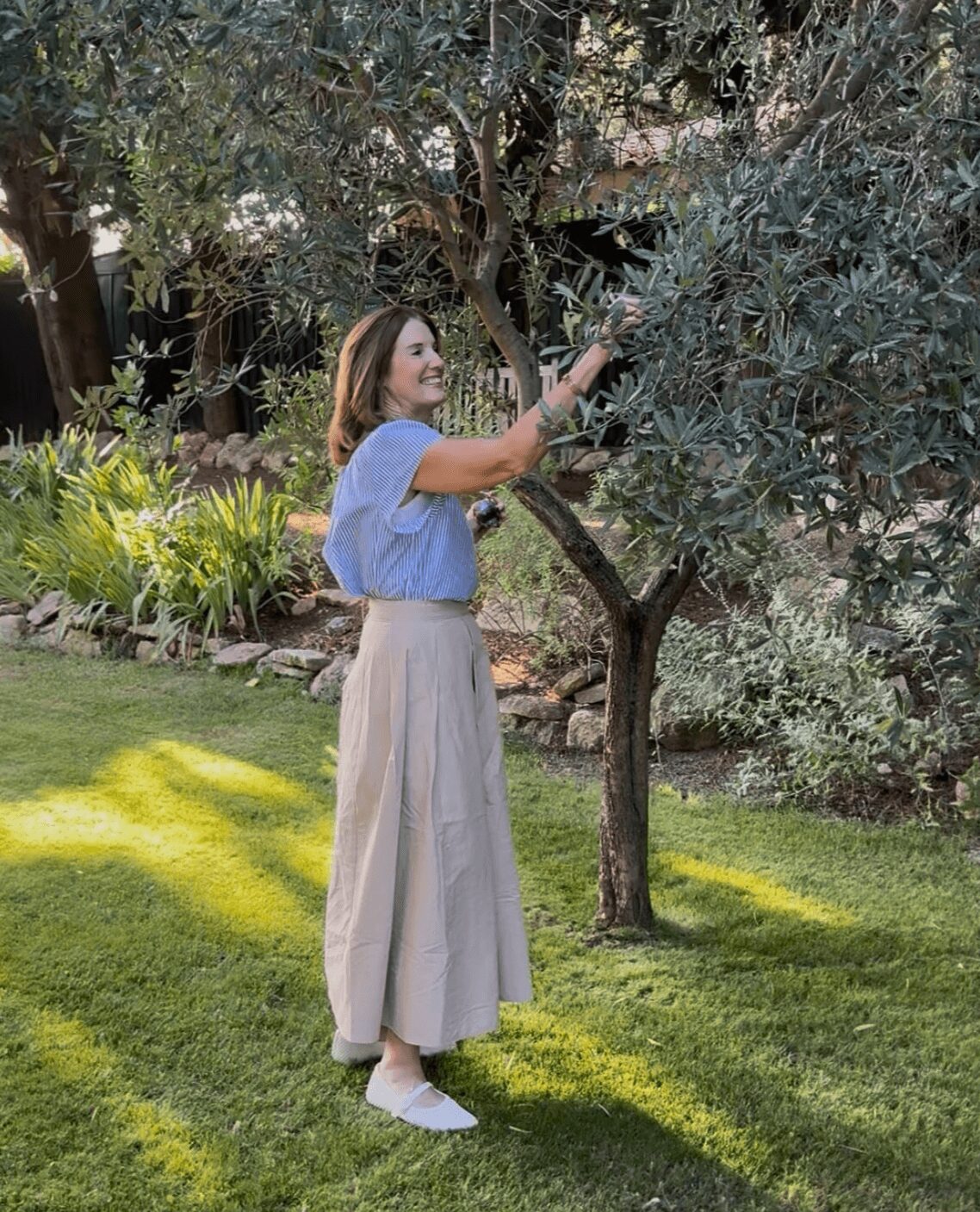 Woman in a blue top and beige wide-leg pants reaching for leaves on a tree in a garden.