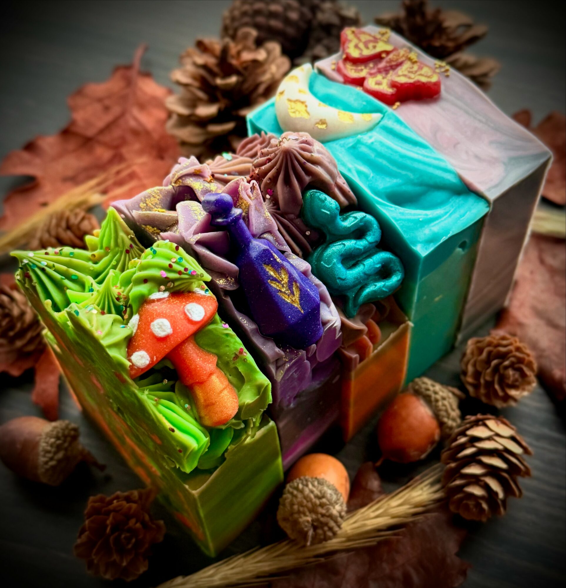 Colorful decorated soap bars with autumn leaves and pinecones around them, on a dark surface.