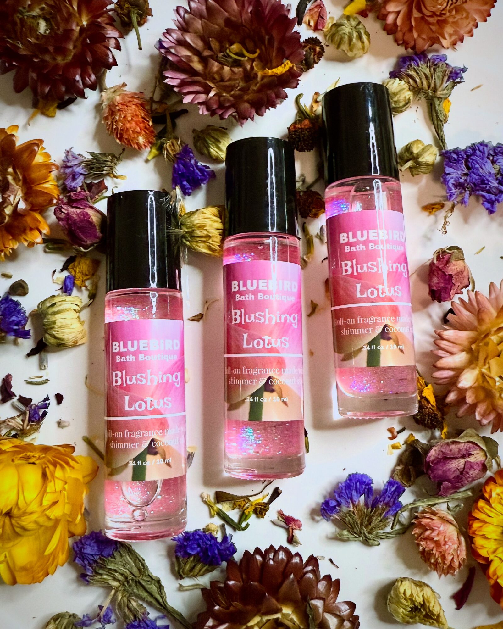 Three pink bottles of body spray surrounded by colorful dried flowers on a white background.