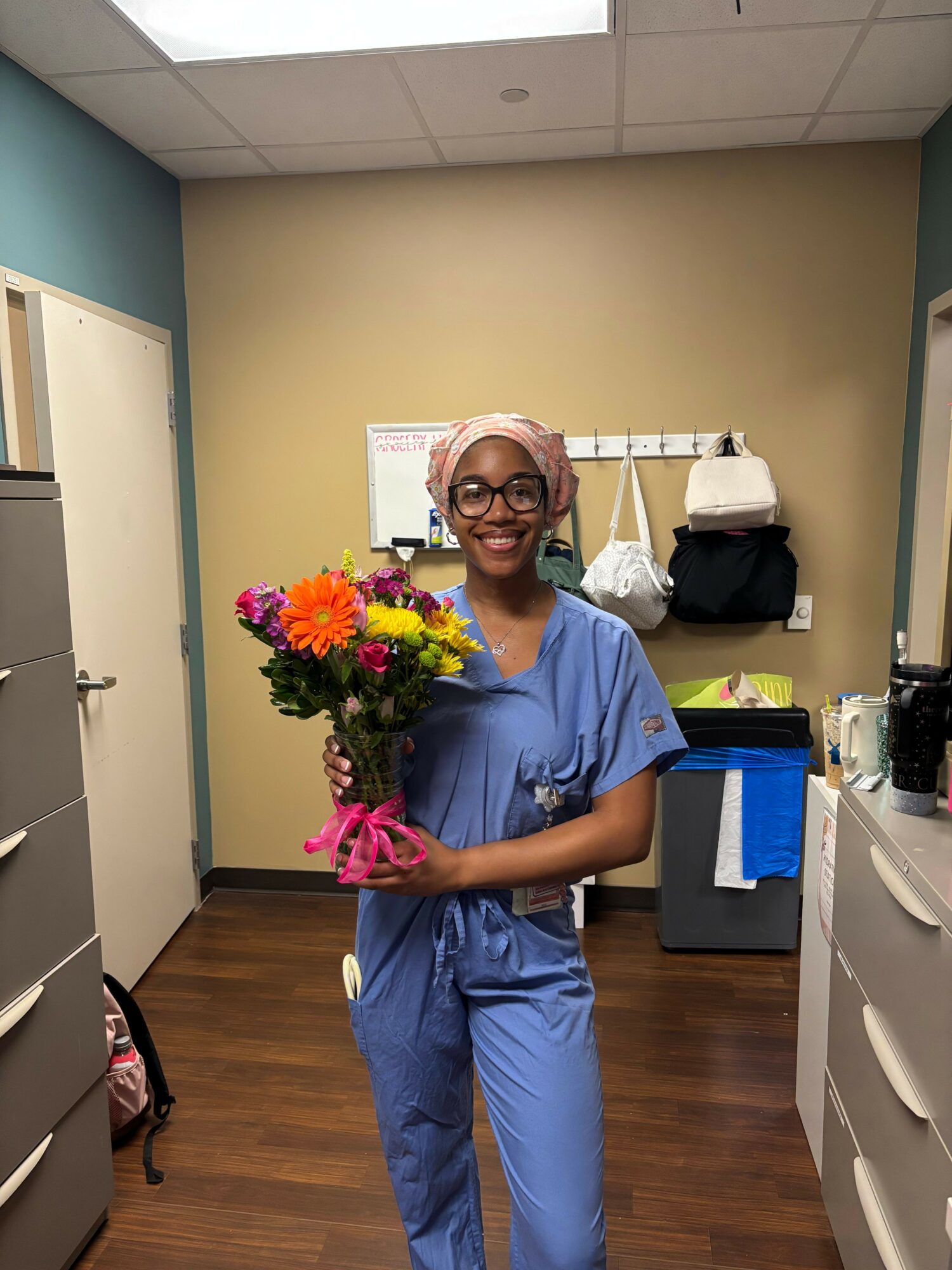 Healthcare worker in scrubs holding a colorful bouquet of flowers in a room with medical supplies and furniture.