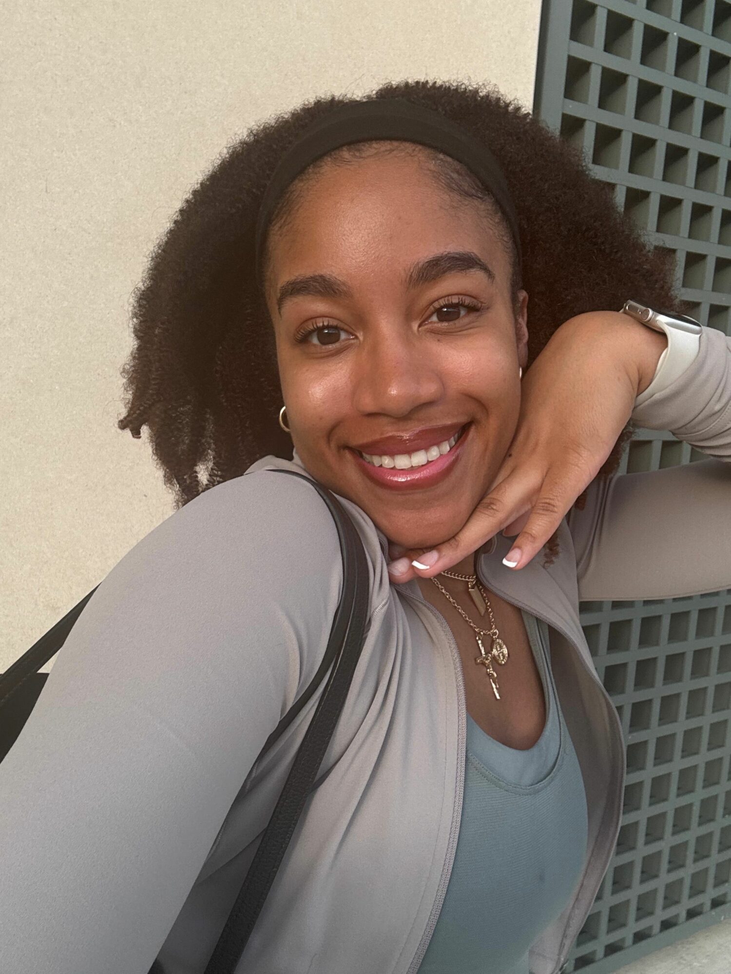 Woman with curly hair smiling, resting her chin on her hand, wearing a gray jacket and a necklace.