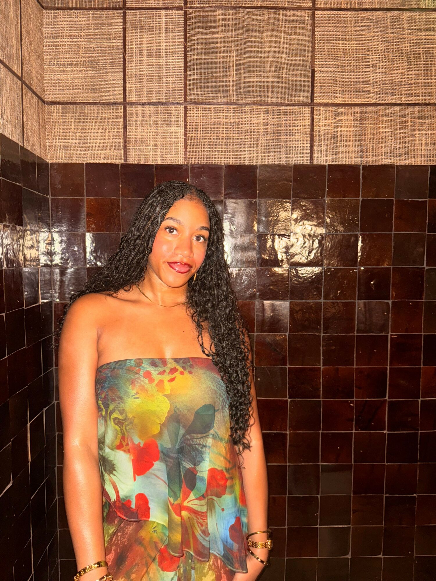 Woman with curly hair in a colorful strapless dress, standing against a tiled wall, smiling at the camera.