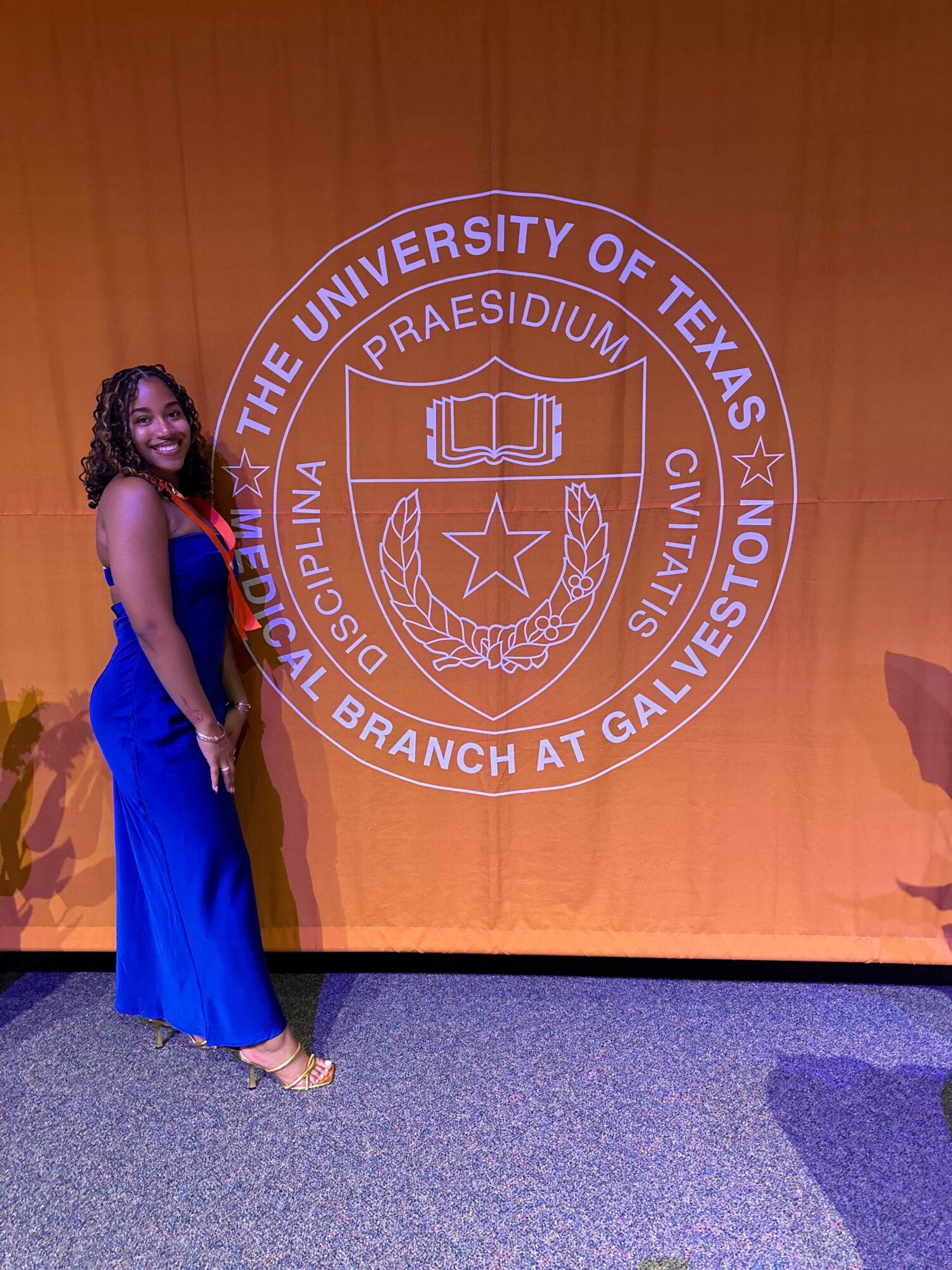 Woman in a blue dress standing on stage with a university seal background, holding a microphone.