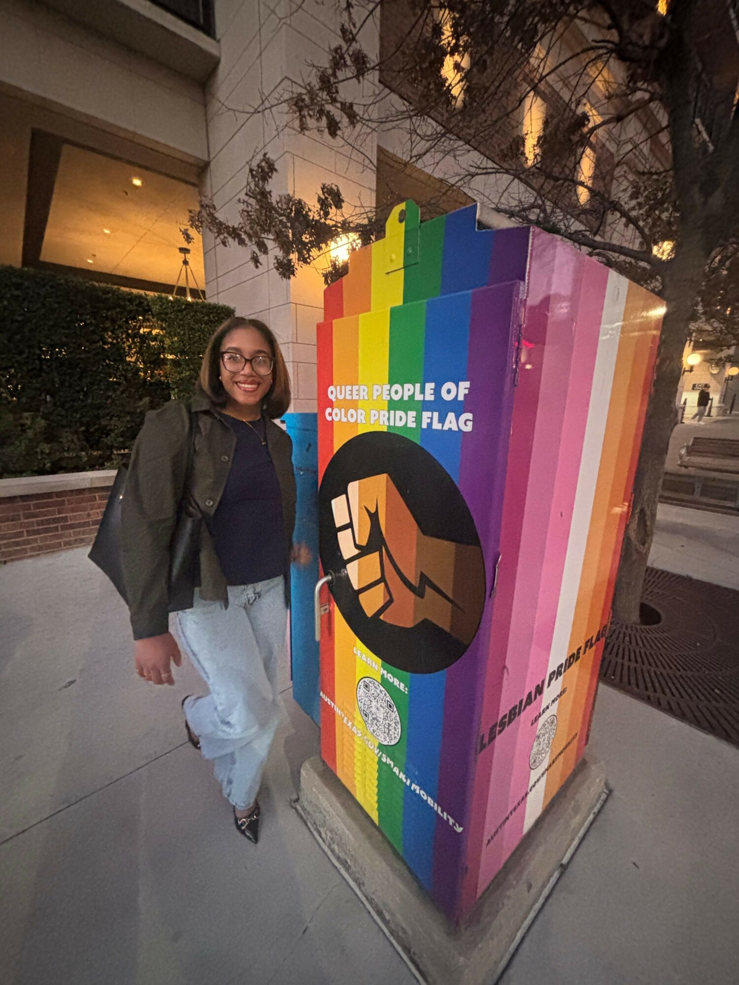 Person standing next to a colorful rainbow flag mural with a fist symbol, outdoors during evening, with trees and buildings nearby.