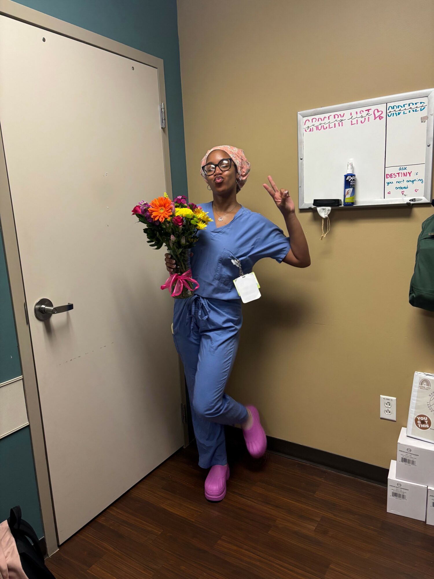 Person in blue scrubs holding a colorful bouquet of flowers, standing in a room with a whiteboard and boxes, smiling.