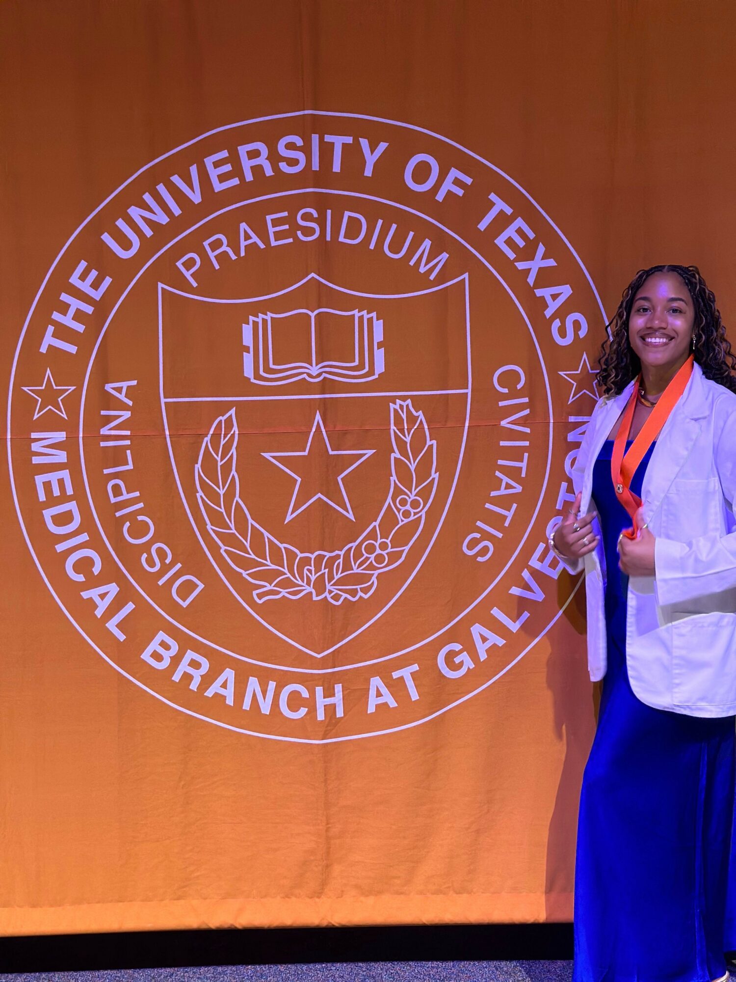 Woman in white coat standing next to a seal of the University of Texas Medical Branch, with orange background.