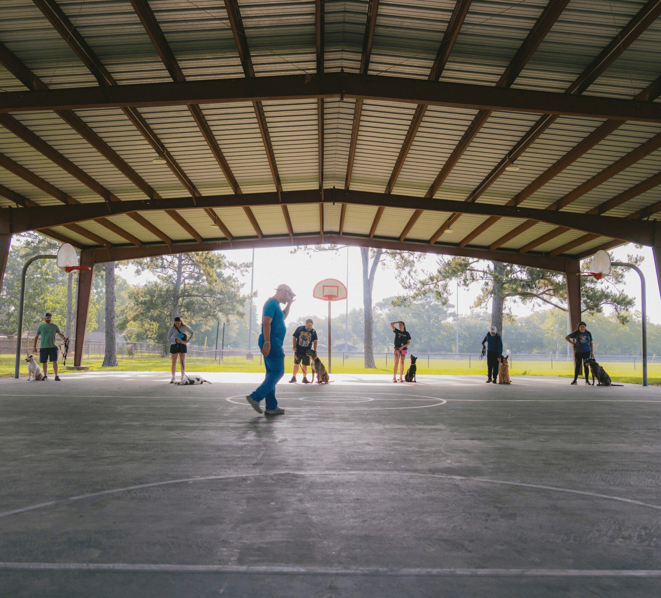 People playing basketball on an outdoor court under a large roof, trees in the background, some children and an adult present.