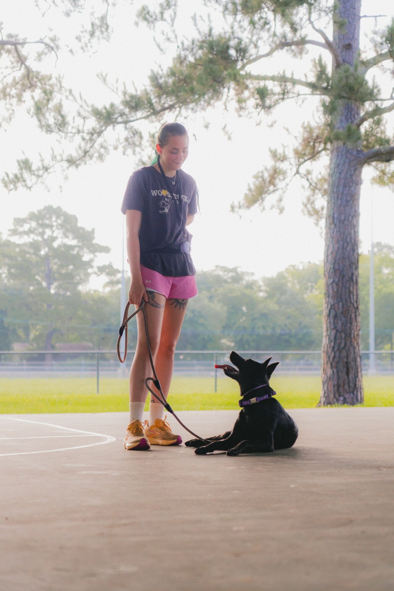 Person standing on a court with a black dog sitting beside them, trees in the background, bright lighting.