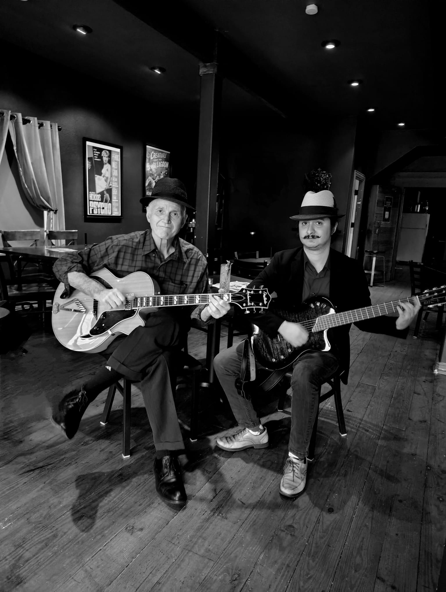 Two people playing guitars sit on chairs in a dimly lit room with framed pictures on the wall.
