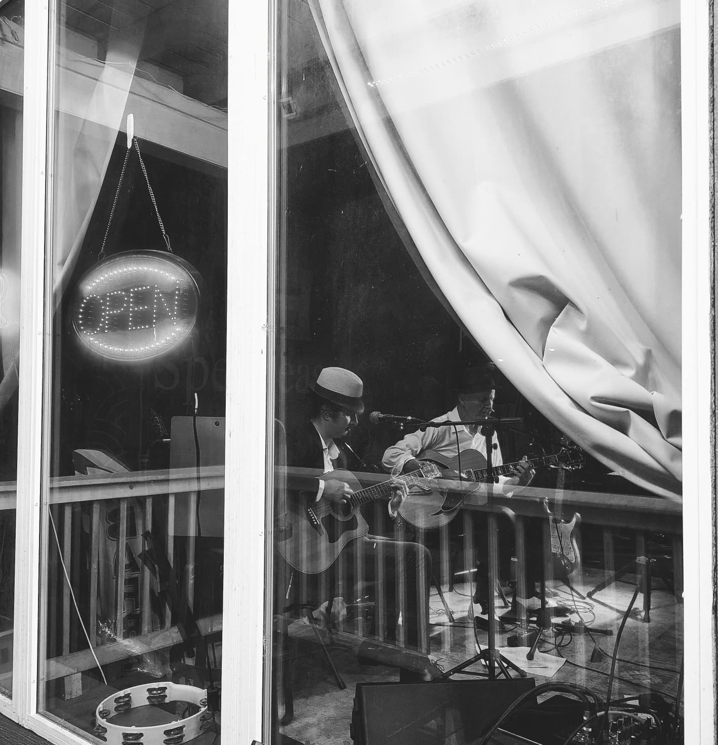 People playing guitars inside a room with a window, curtain, and an illuminated 'OPEN' sign, seen through the glass.