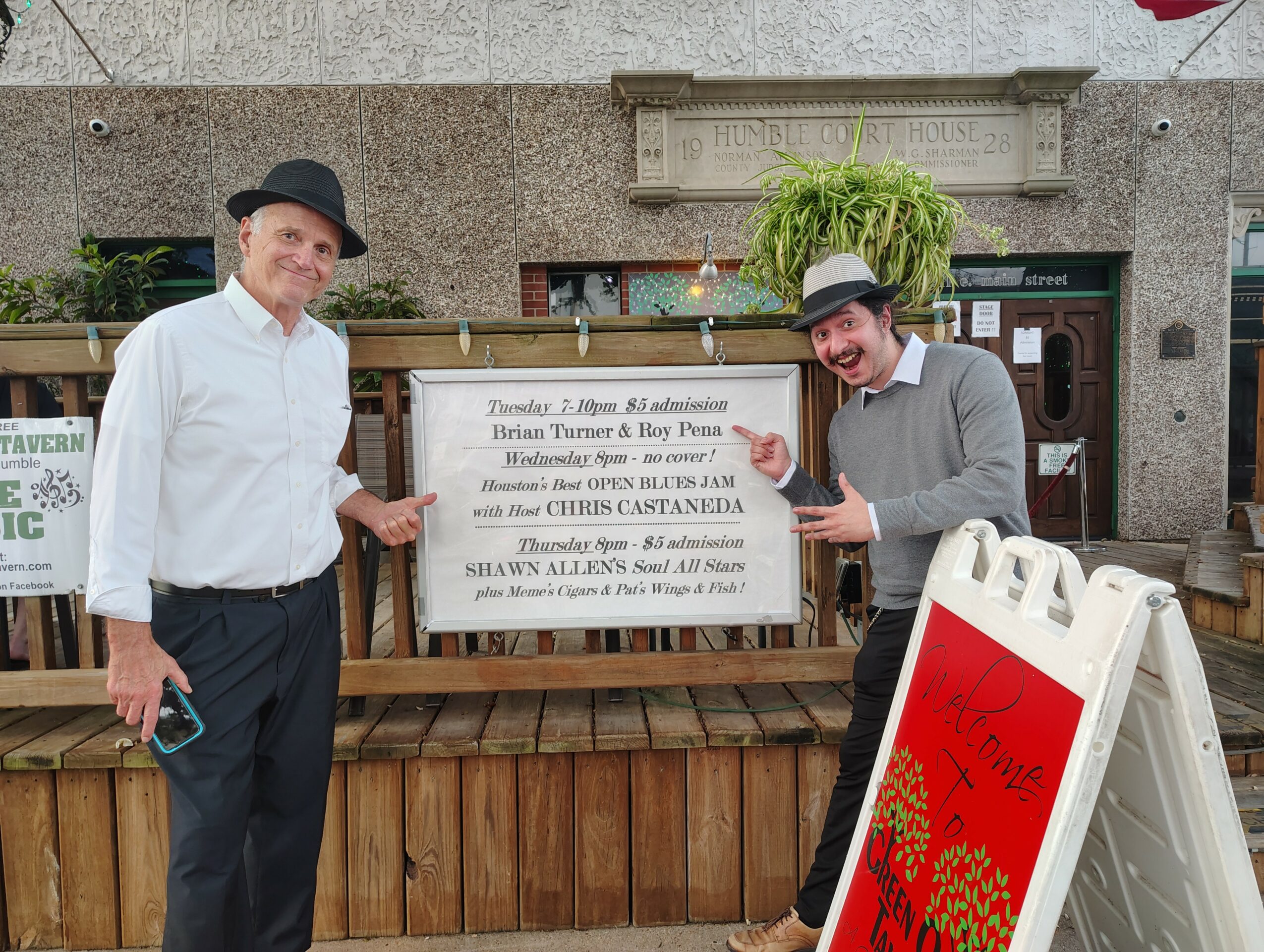 Two men standing in front of a sign, one smiling and wearing a hat, the other laughing and pointing at a red and white signboard, with a building and plants in the background.