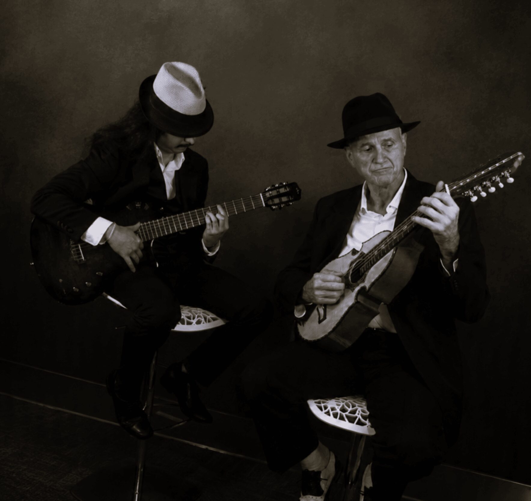Two people playing guitars, one wearing a hat, seated against a dark background.