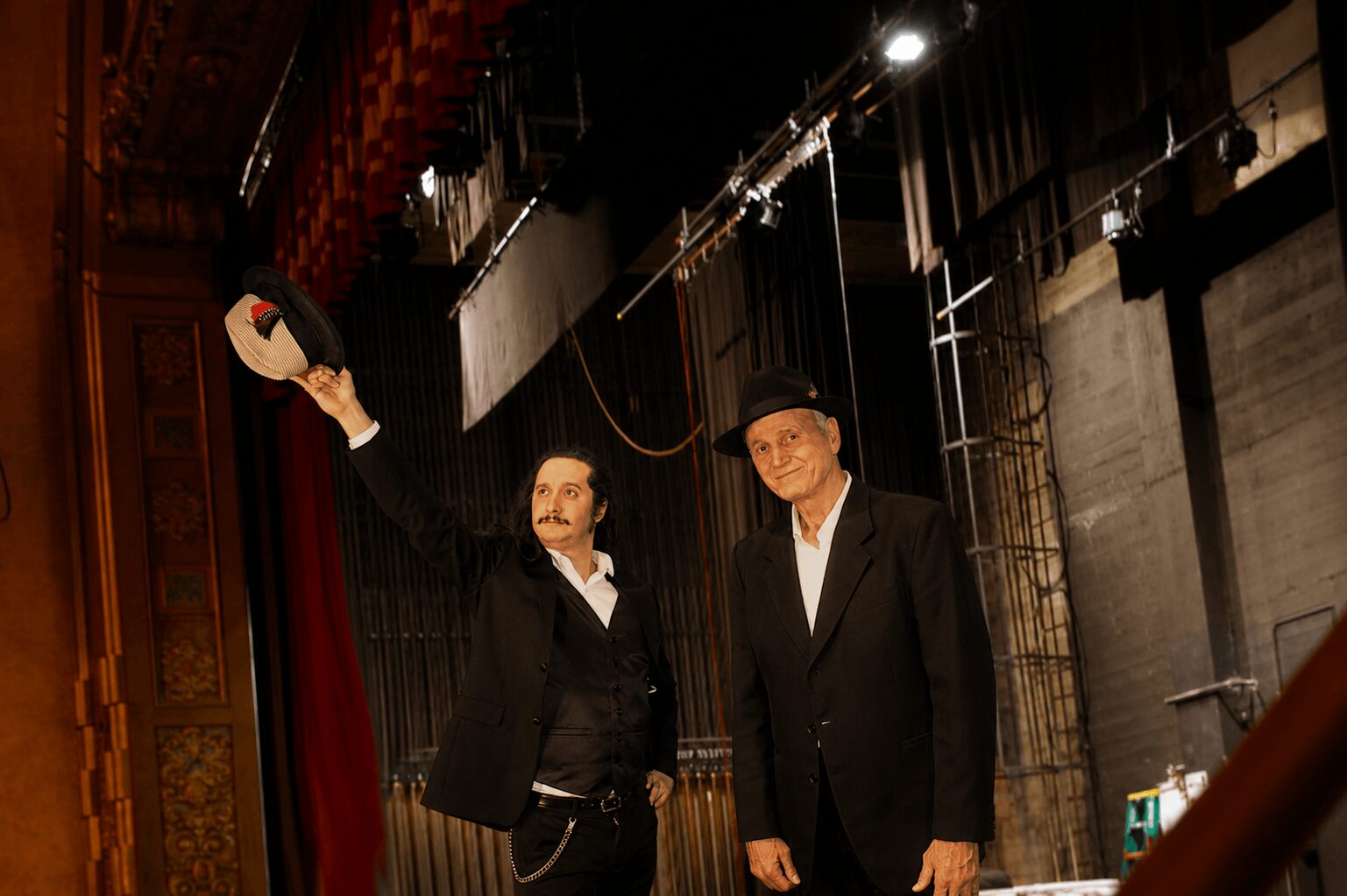 Two men in formal suits and hats standing on stage, one holding a hat in the air, stage lighting above.